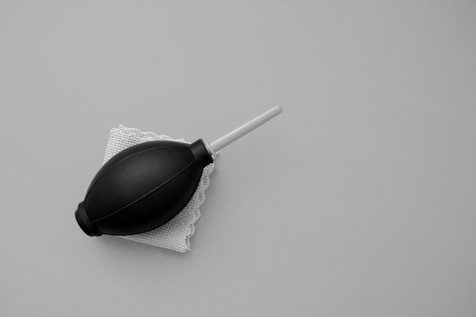 A black rubber bulb syringe with a white plastic nozzle resting on a white cleaning cloth, placed on a smooth, light grey surface. The bulb syringe is positioned with the nozzle pointing towards the u