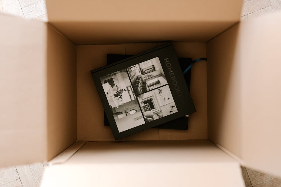 Inside a large cardboard moving box, a black and white hardcover photo book titled 'HOMEBOOK' is visible, showing images of a living room, sofa, fireplace, and decorative interior elements on its cover. The box is placed on a wooden floor near an open doorway, suggesting the packing process during home relocation or furniture transport. The box appears to be partially filled with packing materials and possibly other moving items. The environment indicates a clean, well-lit interior space, typical of a house or apartment being prepared for a move. This scene relates to the services provided by Man and Van Hendon for house removals, including packing and loading processes essential to smooth and efficient moving logistics.