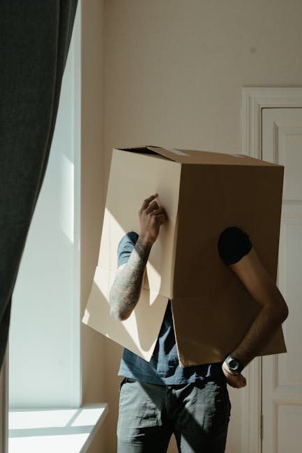 A person wearing a short-sleeved blue shirt and grey trousers is carrying a large cardboard box in a room with white walls and a white door. The individual has tattoos on their left arm and is wearing a black wristwatch on their left wrist. They are holding the box with their left hand, while their right hand is touching their face inside the box. The room is well-lit with natural light coming from a nearby window, partially covered by dark curtains. The scene captures the loading process typical of home relocation or furniture transport, with the person appearing to be in the act of moving boxes as part of a house removal service, such as that offered by Man and Van Hendon. The image emphasizes the handling of packaging materials and the careful movement of belongings during a house move.