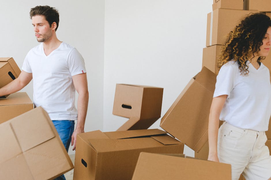 Two individuals involved in a home relocation process indoors, surrounded by numerous cardboard moving boxes in various sizes, some stacked against a plain white wall and others on the floor. The man on the left, dressed in a white t-shirt and blue jeans, is holding and guiding a medium-sized cardboard box, while the woman on the right, wearing a white blouse and light-colored trousers, stands next to a tall stack of boxes, preparing to move or load them. Nearby, a furniture trolley, plastic wrapping, and packing materials are visible, indicating an organized packing and loading process typical of furniture transport and house removals. The environment is well-lit, spacious, and appears to be an interior room ready for the next stage of a professional removal service, such as those provided by Man and Van Hendon, to facilitate efficient packing, transportation, and home relocation activities, supporting the coordination required to avoid common delays in flat removals in Hendon.