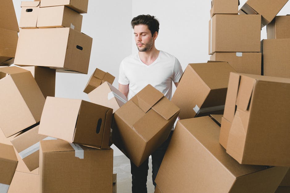 A man with short dark hair and a light beard wearing a white t-shirt is surrounded by numerous cardboard moving boxes in a well-lit indoor space, likely a room in a flat. The boxes vary in size and are stacked and scattered around him, some on the floor and others held in his arms. Many of the boxes have white packing tape applied to secure their flaps. The man appears to be in the process of packing or preparing for a home relocation, with a focused expression on his face. In the background, more boxes are stacked against the wall, indicating an active packing and moving process typical of a residential move. The scene captures the logistical aspect of furniture transport and packing involved in flat removals, areas where Man and Van Hendon offers professional services, ensuring efficient and organized house removals that help avoid delays during the moving process.
