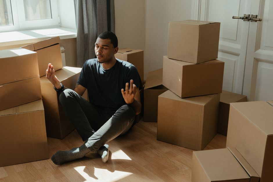 Two individuals involved in a home relocation process indoors, surrounded by numerous cardboard moving boxes in various sizes, some stacked against a plain white wall and others on the floor. The man on the left, dressed in a white t-shirt and blue jeans, is holding and guiding a medium-sized cardboard box, while the woman on the right, wearing a white blouse and light-colored trousers, stands next to a tall stack of boxes, preparing to move or load them. Nearby, a furniture trolley, plastic wrapping, and packing materials are visible, indicating an organized packing and loading process typical of furniture transport and house removals. The environment is well-lit, spacious, and appears to be an interior room ready for the next stage of a professional removal service, such as those provided by Man and Van Hendon, to facilitate efficient packing, transportation, and home relocation activities, supporting the coordination required to avoid common delays in flat removals in Hendon.