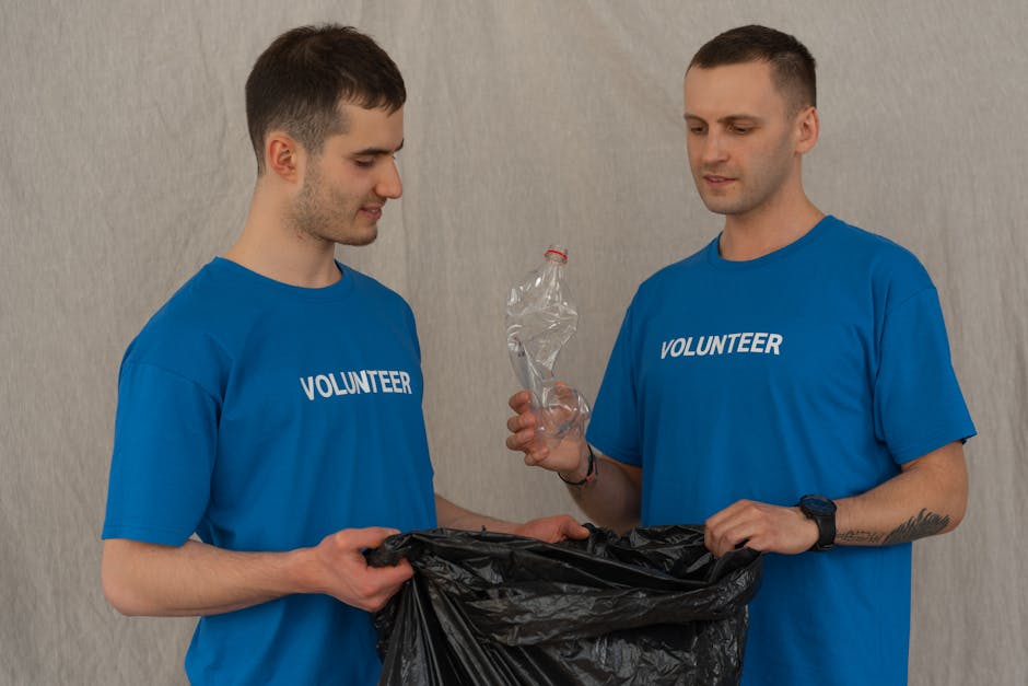 Two male volunteers wearing blue T-shirts with 'VOLUNTEER' printed on the front participate in a home relocation and recycling initiative. The volunteer on the left, with short brown hair, holds a bla