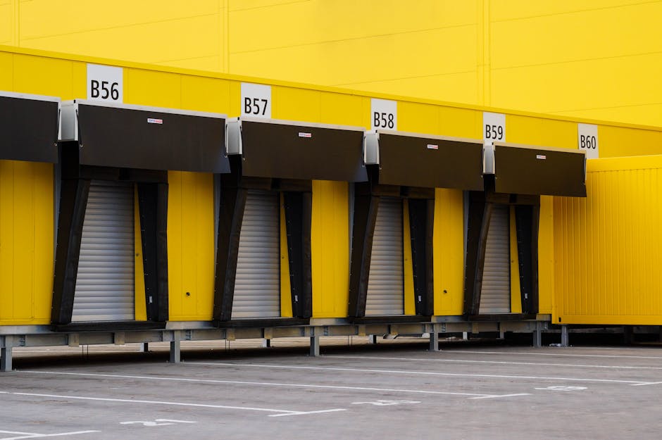 Exterior view of a large yellow industrial building with multiple black loading bay doors labeled B56, B57, B58, B59, and B60, each equipped with black awnings. The doors are closed, with grey roller shutters underneath, and the loading bays are elevated on metal supports. The ground in front of the building is paved with marked parking spaces, and the overall scene appears to be part of a logistics or storage facility suitable for home relocation and furniture transport activities. The image is well-lit with natural daylight, emphasizing the bright yellow facade and organized loading bays, relevant to moving and loading processes managed by a professional removals service like Man and Van Hendon.