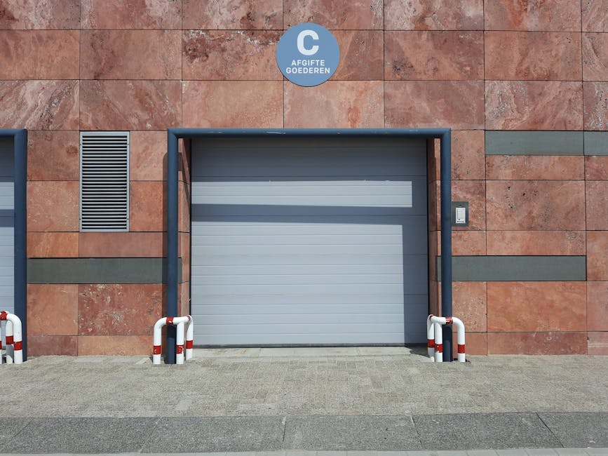 A grey roller shutter door is set into a red brick building exterior with horizontal bands of darker grey bricks. Above the door, a circular blue and white sign indicates 'AFGIFTE GOEDEREN'. On the door’s right side, a small intercom or access keypad is mounted on the wall. The area in front of the door features a paved surface with a small, level concrete pathway. Two white and red striped safety barriers are positioned on either side of the door, preventing vehicle access and ensuring a clear loading space. The setting appears to be an industrial or commercial premises suitable for house removals or furniture transport, supporting loading and unloading activities. The overall environment is well-lit, indicating daytime, and the scene reflects standard measures for safe vehicle and pedestrian movement around loading bays, consistent with Hendon parking suspension rules for removals and loading bays. Man and Van Hendon, a professional removals service, manages the logistics in such areas to facilitate home relocation and furniture transport tasks.