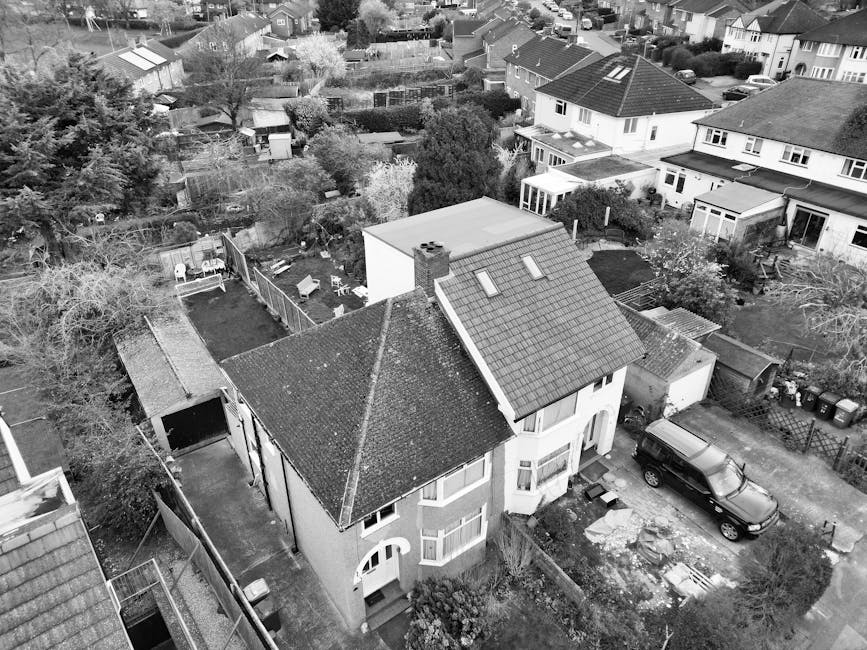 An aerial view of a two-storey detached house with a sloped tiled roof, situated in a residential neighbourhood. The property has a small front garden and a driveway where a black SUV is parked. Outside, a moving process is underway with visible cardboard boxes and packing materials near the entrance. The house is surrounded by other detached and semi-detached homes, with gardens, trees, and fencing separating the properties. In the background, rooftops and backyards of neighboring houses extend into the distance, showing a typical suburban setting. The scene captures part of the loading process for a house removal, with furniture and boxes likely being prepared for transport as part of a home relocation. Man and Van Hendon, specializing in removals, is involved in the logistics of transporting belongings from this property to its new location, supporting the case study of moving from a student flat to a family home in NW4.