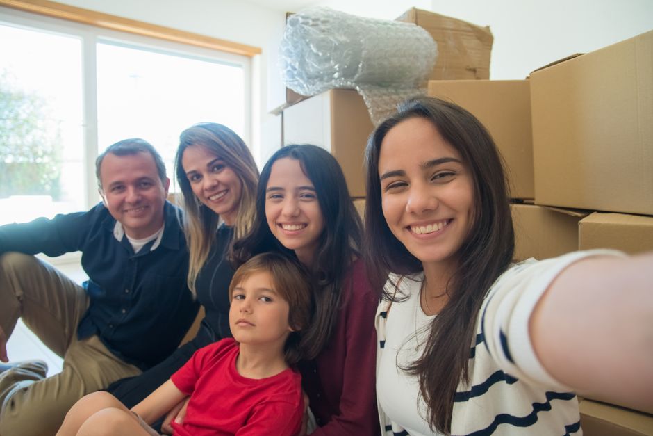 A family of five, including two adult women, one adult man, and two children, sitting together inside a well-lit room with large windows. Behind them are packed moving boxes made of cardboard, some sealed with packing tape, and bubble wrap for fragile items. The furniture includes a white cabinet or wardrobe partially visible in the background. The family members are smiling and appear relaxed, suggesting they are in the process of home relocation. The room has wooden trim around the window and a clean, bright atmosphere. This scene reflects the packing and moving phase of a house removal, with the family posing during the loading process conducted by Man and Van Hendon, a professional removals service in the NW4 area. The image captures the moment of transition from a student flat to a family home, during the packing and furniture transport stage of the house removals.