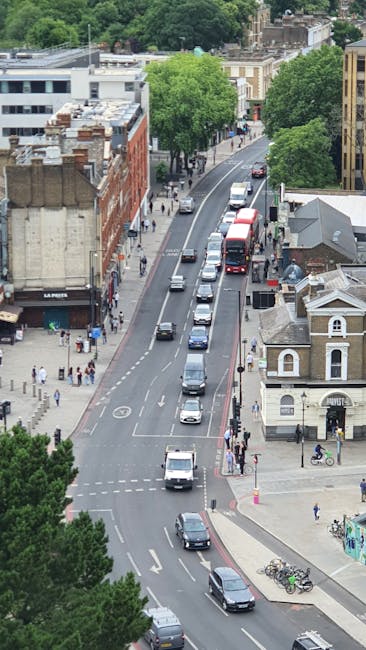 A wide outdoor area in front of Hendon Central station, featuring a modern glass façade building with metal supports, and a spacious paved surface with several people walking, some pushing carts or c