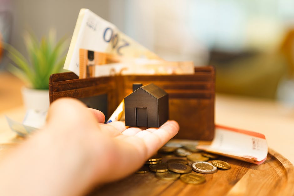 A close-up view of a person's hand reaching into a brown leather wallet positioned on a wooden surface, with some British currency coins scattered nearby. Inside the wallet are folded British banknotes, including a visible £20 note at the top. Adjacent to the wallet, there is a small black miniature house model held in the palm, symbolising home ownership or property. In the blurred background, a potted green plant is visible, adding a natural element to the scene. The setting appears to be indoors, likely a home or office space, with warm lighting overhead. This image relates to house removals and property-related services provided by Man and Van Hendon, highlighting aspects of packing, financial preparation, and moving logistics involved in a home relocation process, consistent with the Hendon removals real cost guide NW4.