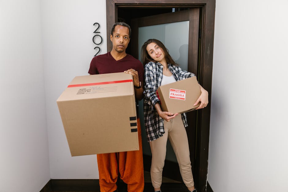 A man and a woman stand indoors next to a doorway, each holding a cardboard box for a home relocation. The man, on the left, is wearing a maroon t-shirt and orange trousers, with a neutral expression, holding a large box with a red and white shipping label. The woman, on the right, is dressed in a checkered shirt over a white top and beige trousers, leaning her head on the door frame with a slight smile, holding a smaller box marked with a 'Fragile' label. Behind them, the door is open, revealing a modern room with white walls and a black floor. The scene takes place during packing and moving preparations, with the visible boxes, packaging materials, and the absence of other furniture indicating the early stages of a house removal process. This image exemplifies the logistics involved in furniture transport and loading during a professional removals service by Man and Van Hendon, supporting the services outlined in the Hendon removals real cost guide NW4.