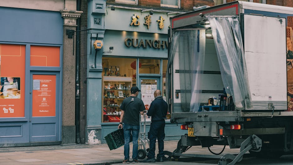 Two men, one wearing a black jacket and the other in a navy coat, stand on the sidewalk in front of a store with Chinese signage and the word 'GUANG' visible. One man holds a plastic crate, while the other has a hand truck nearby. A large moving truck with an open side door, covered partially with clear plastic sheeting, is parked next to the curb. The truck appears ready for loading or unloading, with items possibly inside. The storefront features display shelves with various goods, and the scene is set during daylight with natural lighting. This image captures the process of furniture transport and packing during a home relocation or commercial move, illustrating the logistical tasks involved in urban removals, as provided by [COMPANY_NAME], in alignment with house and office removals services in Hendon.