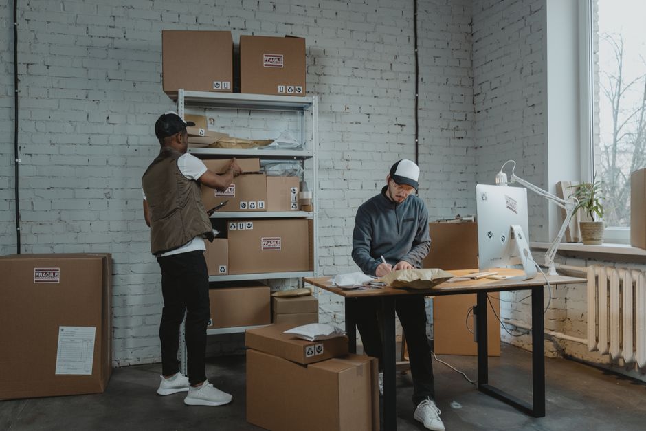 Inside a well-lit room with a white brick wall and a large window, two men are engaged in packing and organising items for a home relocation. One man, dressed in casual clothing with a vest and cap, is standing near a metal shelving unit filled with cardboard boxes labeled with fragile stickers and recycling symbols; he appears to be reaching for or placing a box on the shelf. The second man, wearing a dark hoodie, glasses, and a protective helmet, is seated at a desk, writing on a piece of packing paper surrounded by additional boxes and packing materials. A computer monitor and desk lamp are positioned on the desk, and a potted plant sits by the window, which allows natural light into the space. Several cardboard boxes of various sizes sit on the floor, some sealed with packing tape, indicating an ongoing packing process typical in professional removals. The environment suggests a preparatory stage of furniture transport and packing for an upcoming house move, with necessary equipment like trolleys or straps not visible but implied by the context. Man and Van Hendon is involved in packaging and organising logistics associated with local house removals and furniture transport services.