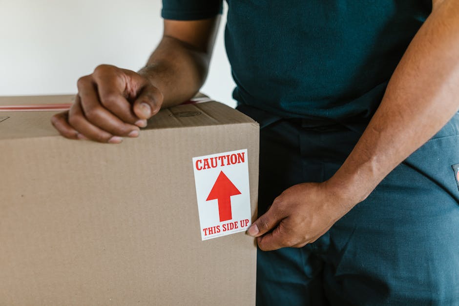 Close-up of a person wearing a dark green shirt and blue trousers, holding a large cardboard box during a house removal. The box has a red and white caution label reading 'CAUTION THIS SIDE UP' with an upward-pointing arrow, indicating careful handling during packing and moving processes. The individual grips the box firmly with both hands, one on the top edge and the other on the side, suggesting an active loading process typical of furniture transport or home relocation. The background shows part of an indoor setting with plain walls, while the person's actions are part of a careful packing and loading sequence carried out by professionals such as Man and Van Hendon, specialists in removals, to ensure safe and efficient furniture transport as part of a house move or relocation project. The image emphasizes proper handling procedures, the importance of clear packing instructions, and the organized logistics involved in domestic removals tailored to the detailed services showcased on the 'RAF Museum move logistics Hendon' page.