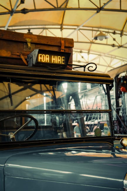 A close-up view of a vintage vehicle's front section, featuring a metal sign mounted on the roof that reads 'FOR HIRE' in illuminated yellow text. The vehicle has a large, clear windshield and a black frame, with parts of the metal body visible in a dark blue or gray color. The setting appears to be an indoor or covered area with a yellow fabric or canvas canopy supported by metal framing visible overhead. The vehicle is part of a move logistics setup, possibly related to house removals or furniture transport, as indicated by the setting reminiscent of a vintage transport or historical-themed environment, with the signage implying its use for hire services related to relocations. The image is captured in good natural lighting, highlighting the polished surface of the vehicle and the mounting details of the sign.