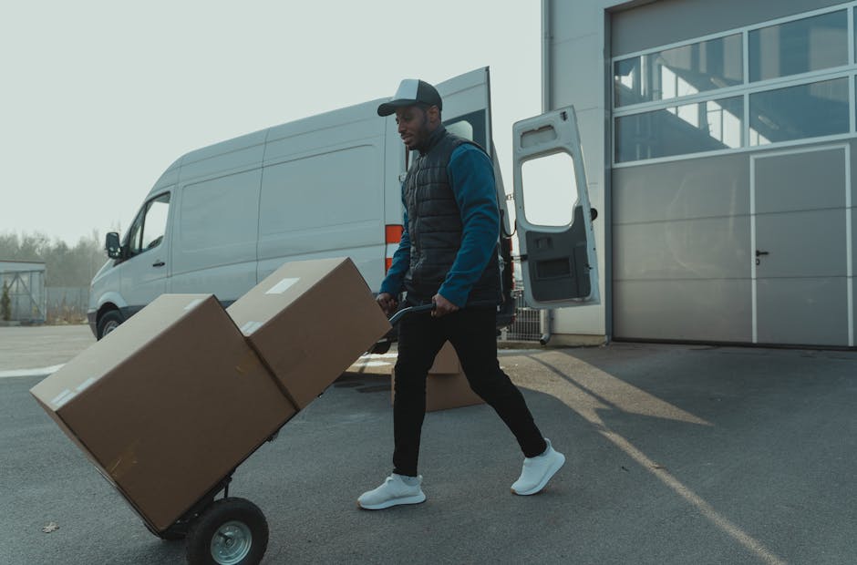 A man in casual outdoor clothing, including a dark baseball cap, blue jacket, and white sneakers, is pushing a hand trolley loaded with two large cardboard boxes towards an open back door of a warehouse or storage facility. The trolley has two black wheels and a metal frame supporting the boxes, which are secured with packing straps. Behind him is a white panel van parked on a paved surface, with its side door open, revealing empty interior space for furniture transport or home relocation. The environment appears to be an outdoor loading area, with a modern building featuring large glass windows to the right, and a partly cloudy sky overhead. The scene captures a moment during a packing and moving process, illustrating the logistics involved in professional removals, with the van positioned to facilitate efficient loading of household items.