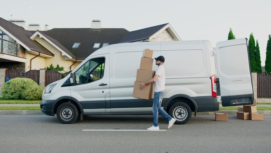 A man wearing a white T-shirt, dark cap, jeans, and white sneakers is shown unloading three medium-sized cardboard boxes from the side of a white commercial van parked on a residential street in Hendon. The man is carrying one box in his arms while the other two are stacked on top of each other, held against his body. Additional cardboard boxes are placed on the pavement near the van's open side door. The van is positioned parallel to the curb, with its sliding door open to facilitate the loading process. In the background, there are modern houses with pitched roofs, brown wooden fences, well-maintained lawns, and tall green trees. The scene is illuminated by natural daylight, suggesting daytime hours. This image, associated with Man and Van Hendon, illustrates a professional home relocation or furniture transport service involving packing and loading activities as part of a house removals process.