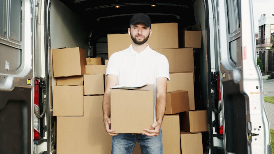 A man in casual outdoor clothing, including a dark baseball cap, blue jacket, and white sneakers, is pushing a hand trolley loaded with two large cardboard boxes towards an open back door of a warehouse or storage facility. The trolley has two black wheels and a metal frame supporting the boxes, which are secured with packing straps. Behind him is a white panel van parked on a paved surface, with its side door open, revealing empty interior space for furniture transport or home relocation. The environment appears to be an outdoor loading area, with a modern building featuring large glass windows to the right, and a partly cloudy sky overhead. The scene captures a moment during a packing and moving process, illustrating the logistics involved in professional removals, with the van positioned to facilitate efficient loading of household items.