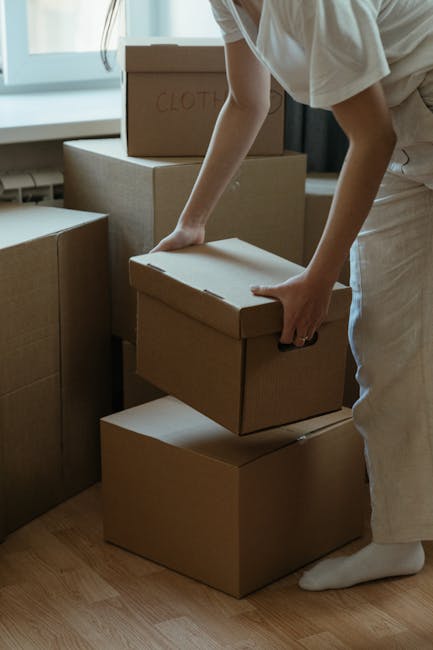 A person wearing white clothing is seen handling a cardboard box during a home relocation, with several other boxes stacked nearby in a well-lit room. The individual is grasping the box with both hands, preparing to move or position it as part of the packing and moving process. The boxes are made of corrugated cardboard, some with handwritten labels, and are arranged on the hardwood floor close to a window that provides natural light. Additional packing materials, such as bubble wrap or packing paper, are not visible, indicating a focus on the furniture transport and packaging by [COMPANY_NAME]. This scene illustrates the careful handling involved in house removals and furniture transport, with a clear emphasis on safe lifting techniques. The setting suggests an organized space ready for a move, with the boxes likely containing personal belongings or household items, aligned with the service offered in the West Hendon Broadway area, emphasizing the logistical aspects of both packing and loading during a house removal.