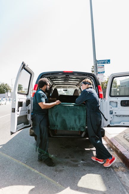 Two movers from Man and Van Hendon are engaged in a home relocation process, standing at the open rear doors of a white van on a street in West Hendon Broadway. The worker on the left, with medium skin tone and dark hair, is holding the edge of a large, dark green upholstered piece of furniture, possibly a sofa or armchair, which is partially covered with packaging material for protection. The other worker, with light skin and wearing glasses, a dark jacket, and red sneakers, is assisting by steadying or lifting the furniture. Inside the van, visible boxes and packing materials such as cardboard and plastic wrap are stacked, indicating a packing and moving activity. The scene occurs in bright daylight, with a clear sky and a street sign visible overhead, and the surroundings including sidewalks and parked vehicles, all typical of a residential or commercial area. This image exemplifies the logistical steps involved in furniture transport during a professional removals service provided by Man and Van Hendon, especially in narrow access situations such as West Hendon Broadway.