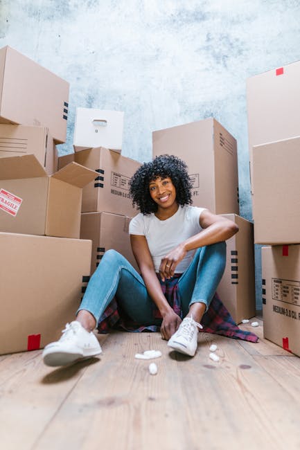 A person wearing white clothing is seen handling a cardboard box during a home relocation, with several other boxes stacked nearby in a well-lit room. The individual is grasping the box with both hands, preparing to move or position it as part of the packing and moving process. The boxes are made of corrugated cardboard, some with handwritten labels, and are arranged on the hardwood floor close to a window that provides natural light. Additional packing materials, such as bubble wrap or packing paper, are not visible, indicating a focus on the furniture transport and packaging by [COMPANY_NAME]. This scene illustrates the careful handling involved in house removals and furniture transport, with a clear emphasis on safe lifting techniques. The setting suggests an organized space ready for a move, with the boxes likely containing personal belongings or household items, aligned with the service offered in the West Hendon Broadway area, emphasizing the logistical aspects of both packing and loading during a house removal.