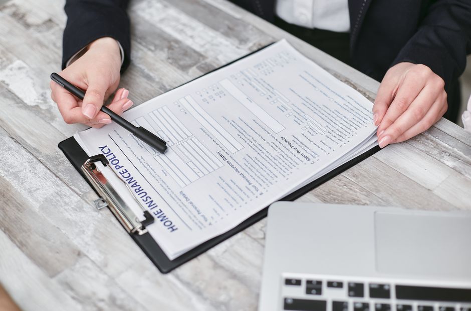 Close-up image of a person in a beige blazer holding a black clipboard with a completed insurance form, using a black pen to point at specific sections. The individual’s left hand supports the lower