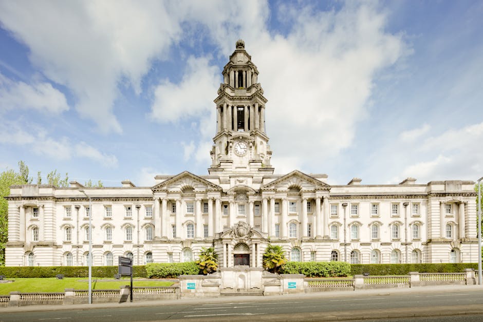 A large historic stone building with a central clock tower featuring multiple levels and ornate architectural details, situated along a paved street. In the foreground, there are well-maintained green hedges and a small garden bed with potted plants. The building is illuminated by natural daylight, with a partly cloudy sky overhead. This property is likely being prepared for home relocation or furniture transport, as part of a moving process. Visible in the vicinity are street signs, and the building's façade includes symmetrical windows and decorative columns. The setting suggests a professional removals service, such as Man and Van Hendon, involved in packing, loading, and transporting household items, possibly in preparation for a move or clearance, with equipment like trolleys or blankets not visible but implied in the context of furniture transport and house removals.